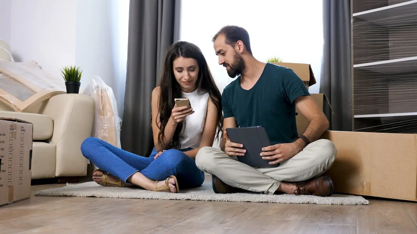 Ein schönes junges Paar, das gerade in eine neue Wohnung eingezogen ist, sitzt auf dem Boden und sucht online nach Möbeln.