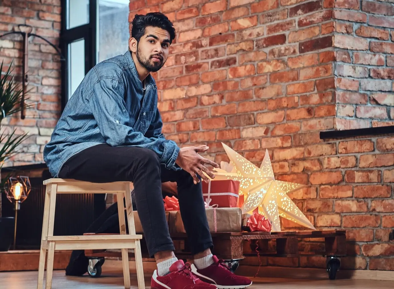 Indischer bärtiger Hipster-Mann mit Tätowierungen am Arm in einem Loftzimmer mit Weihnachtsdekoration.