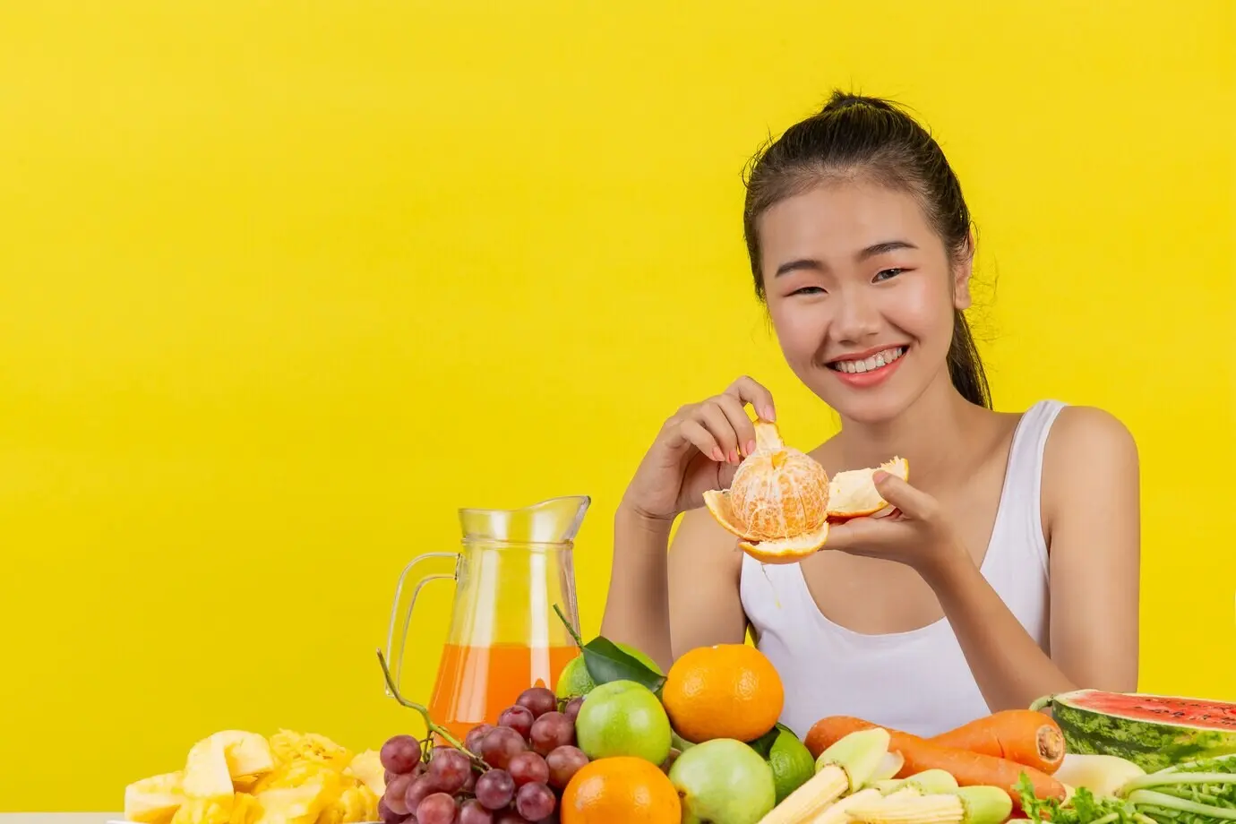 Eine asiatische Frau in einem weißen Tanktop. Ich schäle Orangenschale, und der Tisch ist voller verschiedener Obstsorten.